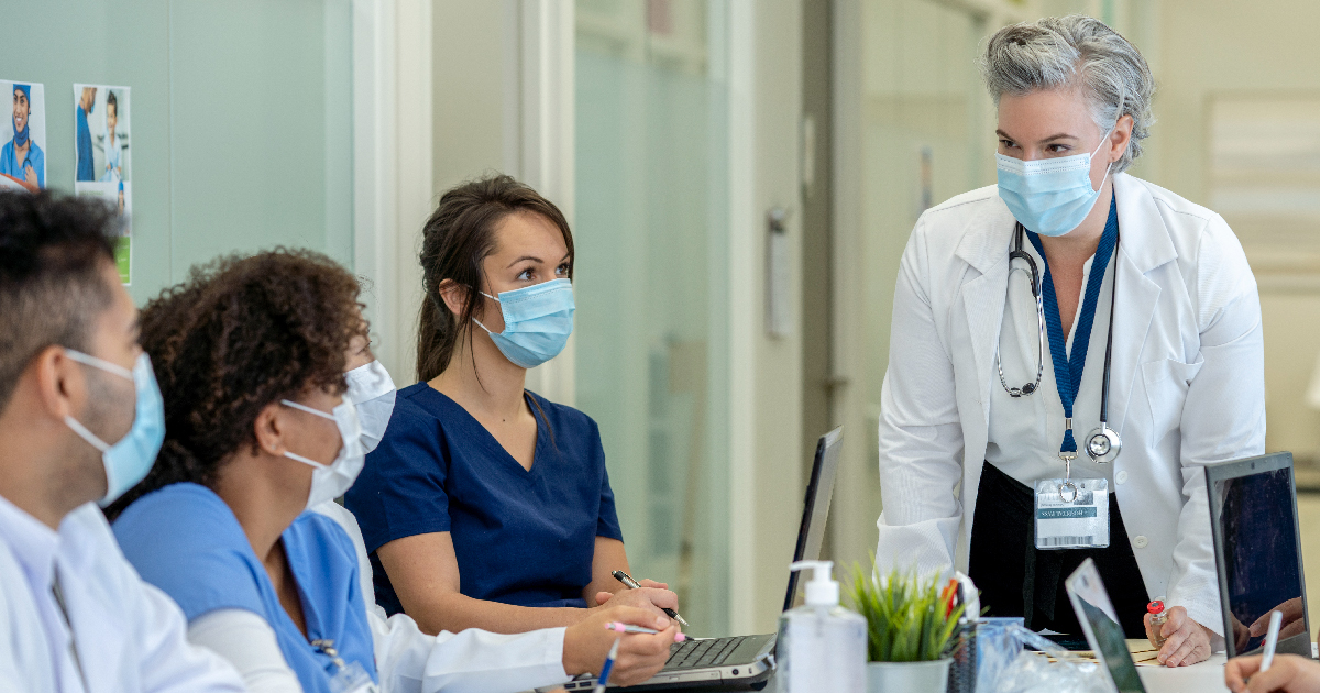 A standing female doctor lectures to three seated med students.