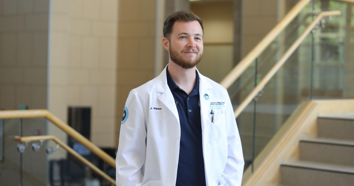 A Doctor of Osteopathy from Kansas College of Osteopathic Medicine wears a lab coat and smiles while looking off-camera.