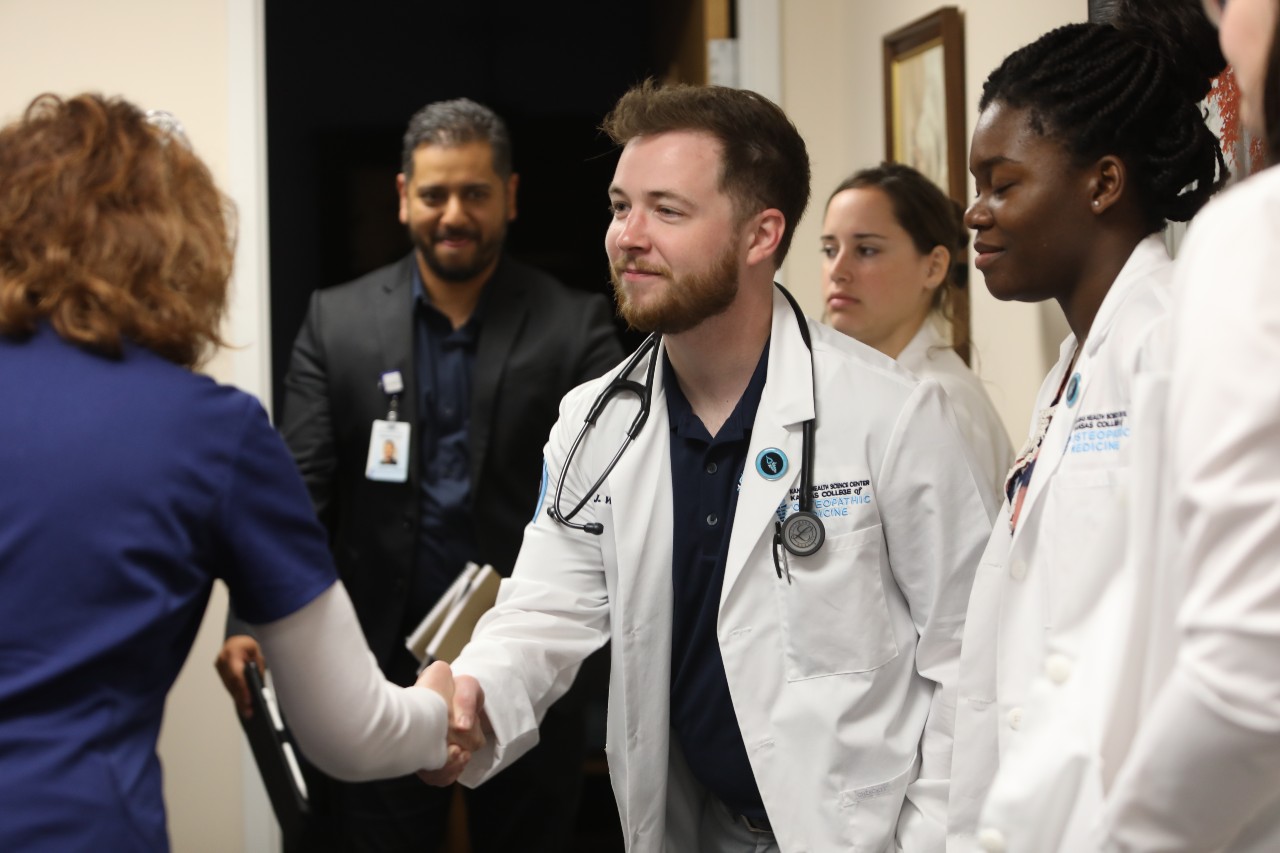 Doctor in white coat shakes hands with one in blue as others look on.