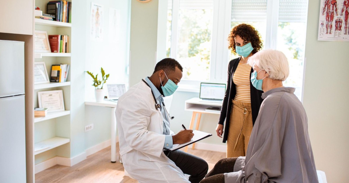 A doctor writes on a clipboard while sitting with a patient in a doctor's office.