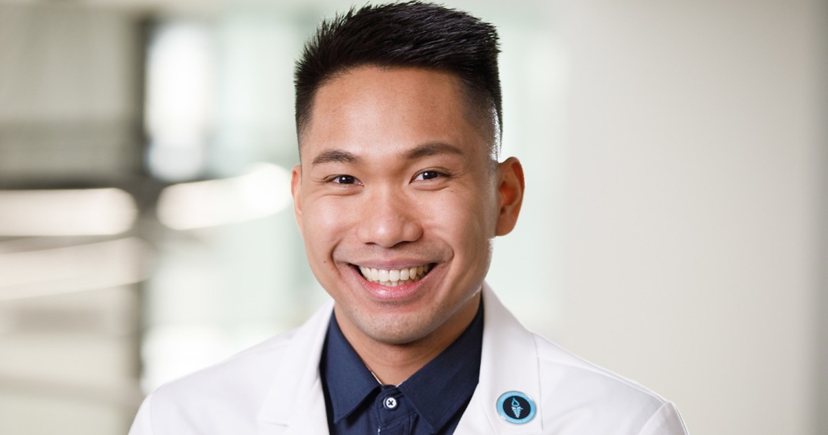 A young Asian man wearing a white lab coat looks at the camera and smiles.