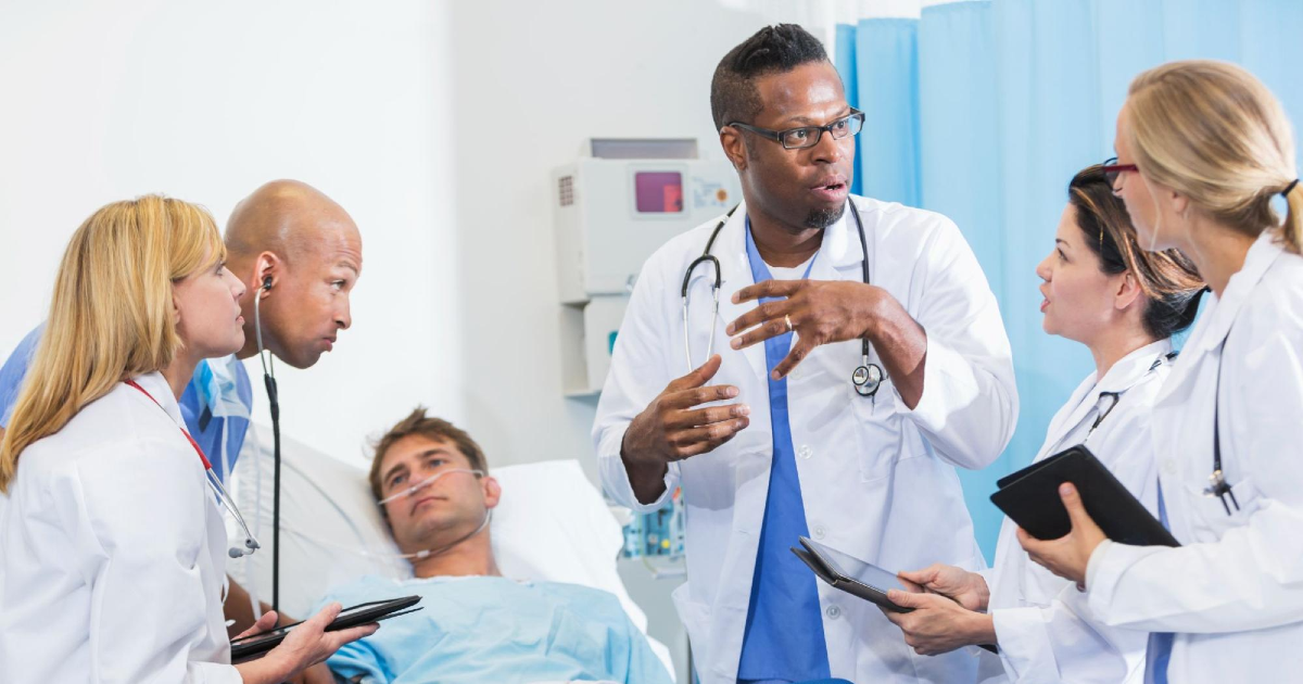 Doctors surrounding a patient's bed listen while another doctor address them and gestures while speaking.