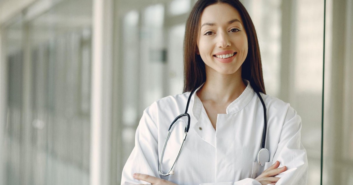 A brunette woman wearing a white lab coat with a stethoscope around her neck crosses her arms and smiles.