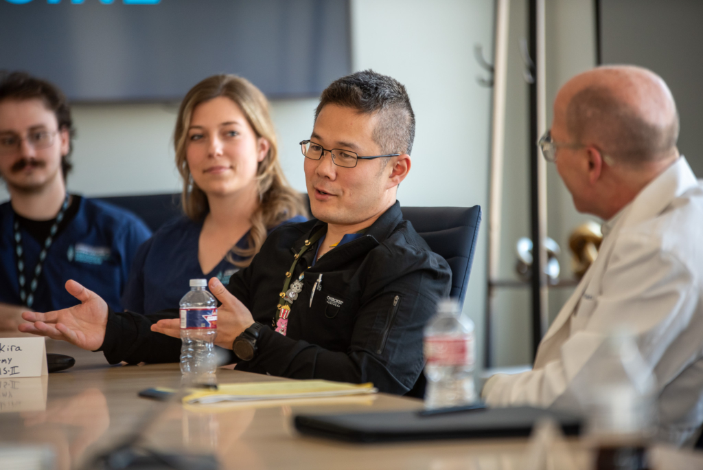 A man gestures while speaking to a group of listeners seated around him at a conference table.