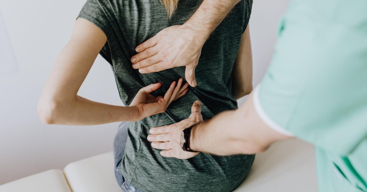 In close-up, a doctor places his hands on a patient's back to examine the patient.