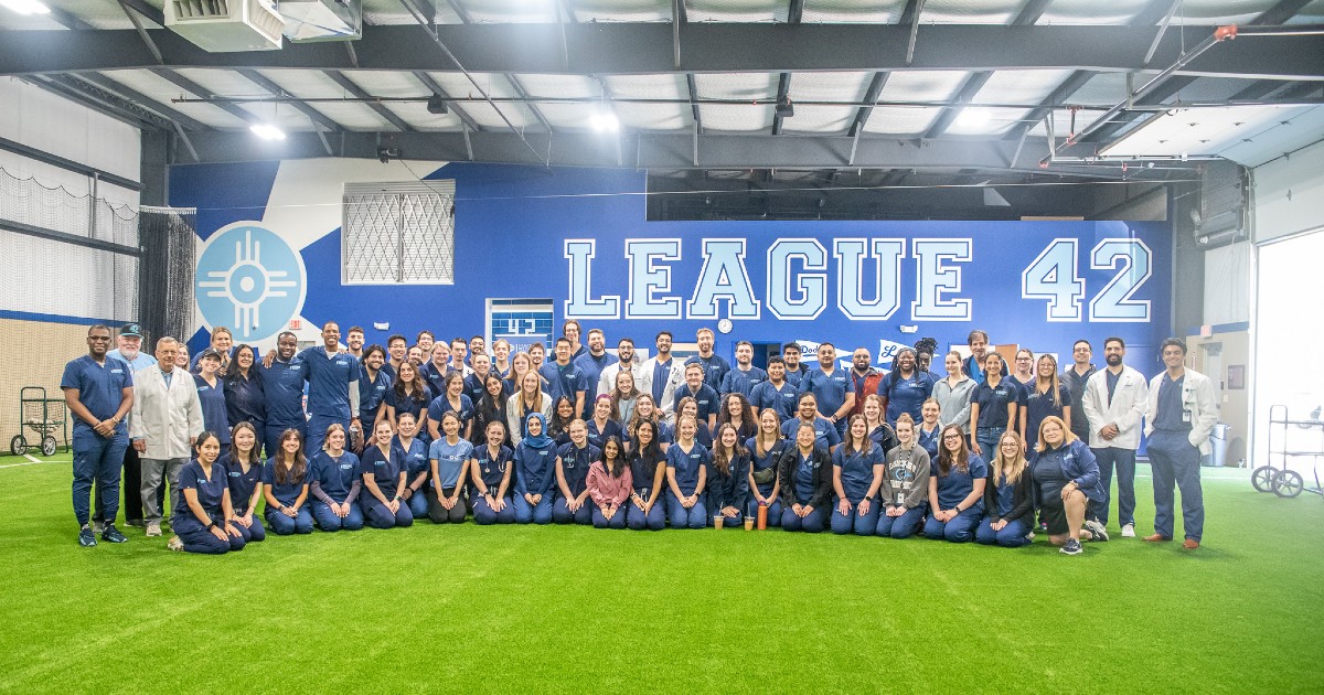 KansasCOM students and members of League 42 pose together in a group photo on an indoor field.